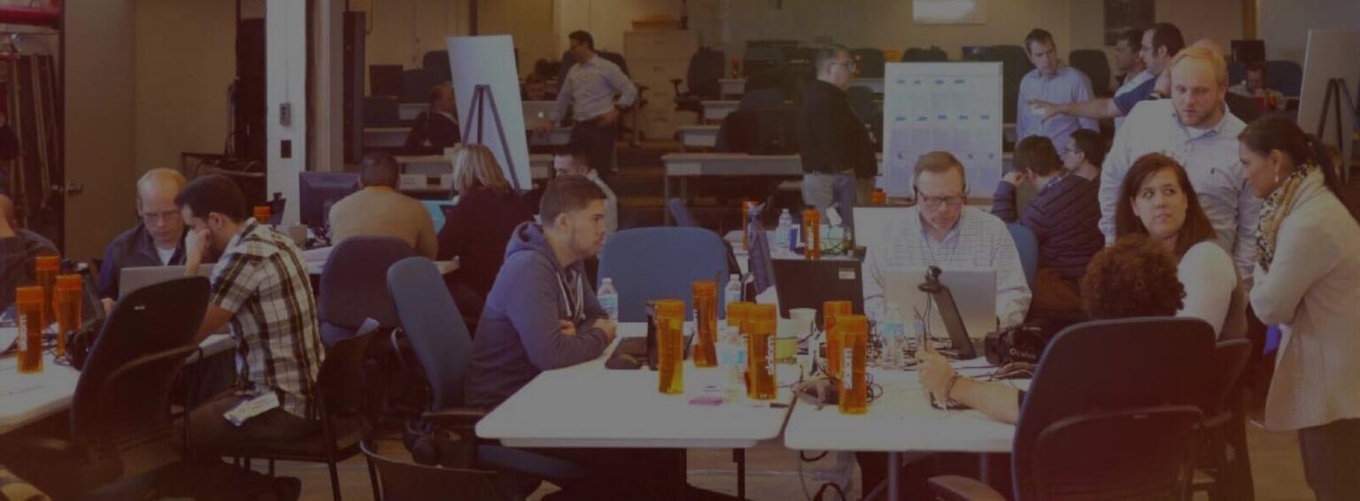 Multiple people sitting at several tables in a large room, white board charts in the background.