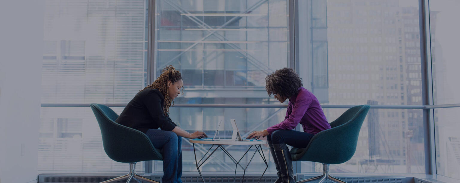 Two women facing each other working on laptops in front of large glass window.