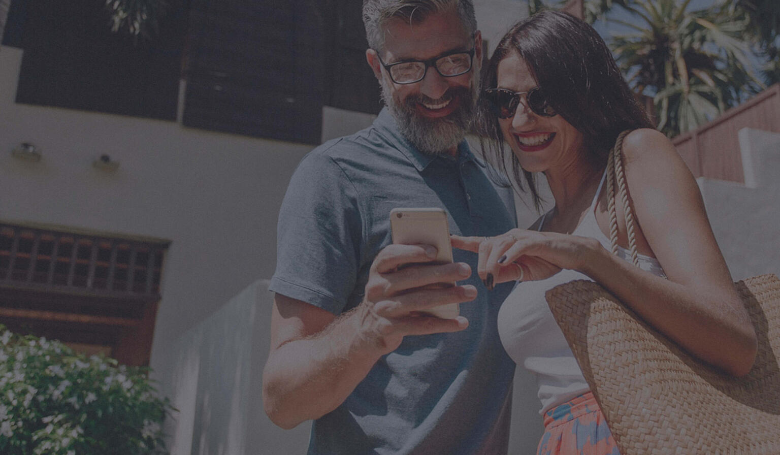 Man and woman in front of tropical house and palm trees pointing at cell phone.