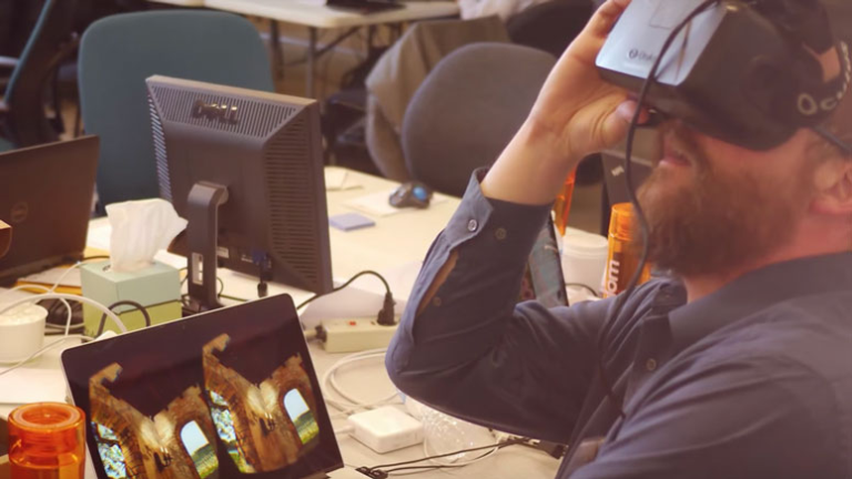 Bearded man sitting at a table, with right hand holding the VR headset he's wearing.